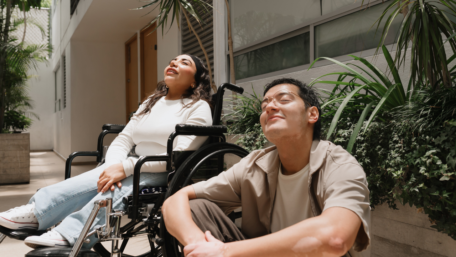 A woman in a wheelchair is sitting with a man next to her. They are pointing their faces towards the sun with their eyes closed, quietly thinking. 