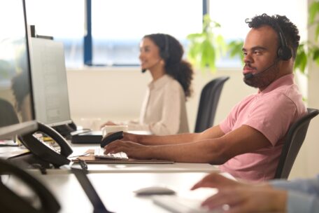 a man with down syndrome is helping people over the phone at his place of work. He is wearing a pink top and headphones while typing something. 