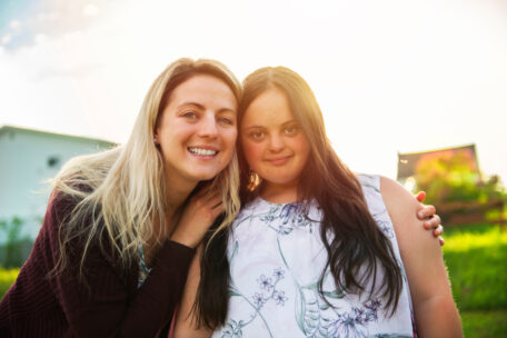 a young woman with trisomie 21 is hugged by her sister in a park. They have just submitted a change of details for SDA