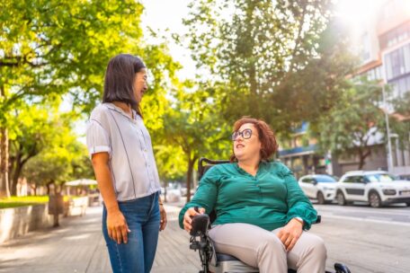 a woman in a wheelchair looks up at her friend as they walk down a tree lined street. 