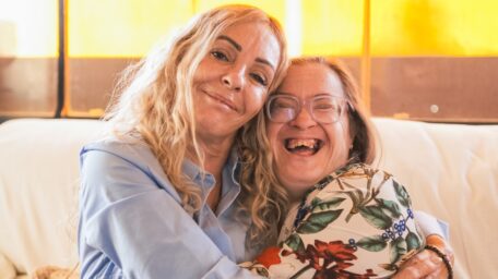 A woman with a disability hugs her sister on the couch, they are considering a change of details to change from plan managed to agency managed SDA