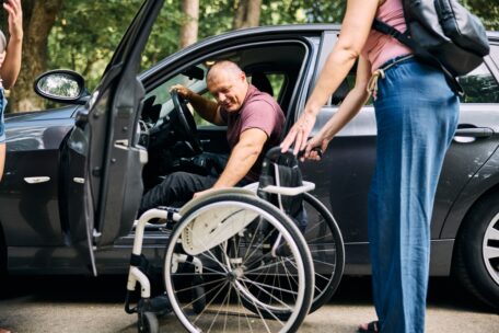 a mans carer helps him by bringing his wheelchair to the drivers door