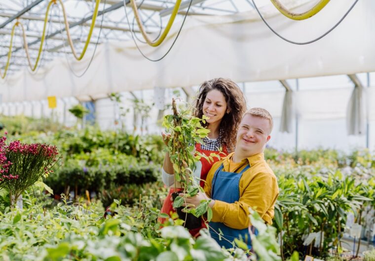 A young man with down syndrome stands with his support worker in a plant nursery. They are considering the Participant Service Guarantee and how nothing is completed on time.