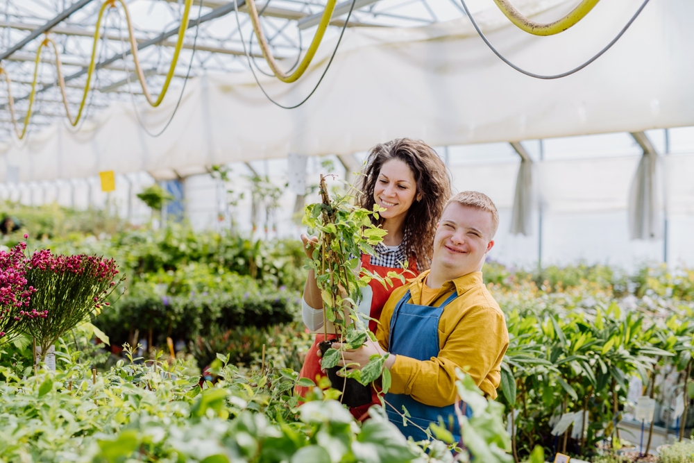 A young man with down syndrome stands with his support worker in a plant nursery. They are considering the Participant Service Guarantee and how nothing is completed on time.