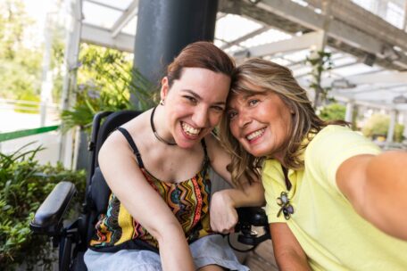 A woman with cerebral palsy takes a selfie with her support worker. They are sitting in a covered area with plants everywhere. 