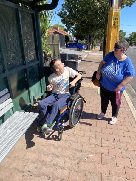 Ryan and his mum look at each other while at a bus top. Ryan is sitting in his wheelchair while his mum stands. 