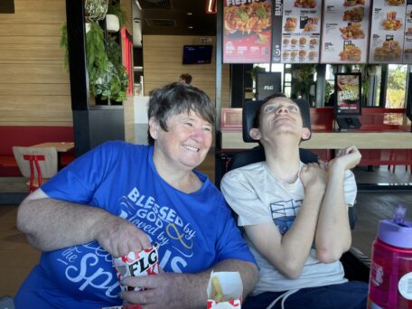Ryan and his mum sit side by side at a restaurant. Sheree is leaning over to Ryan while he makes a happy face. 